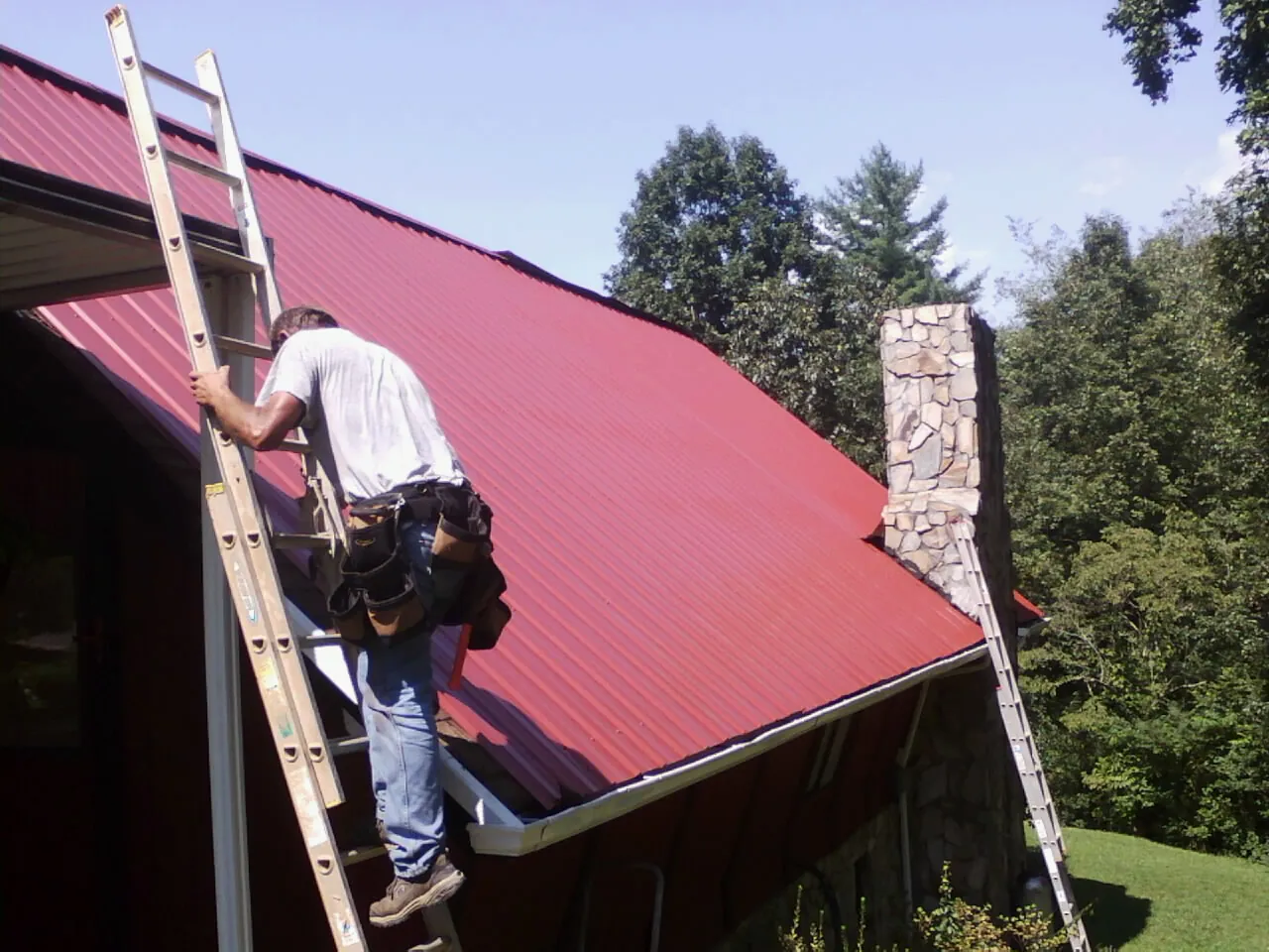 Roofer on a ladder beside a red metal roof and stone chimney — Soffit & Fascia Repair by Roofing Brothers in Kerhonkson